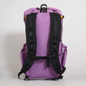 The back view of a purple (mulberry) backpack with black padded shoulder straps and side pockets. Made from X-Pac VX21 fabric. Shown against a light gray background.