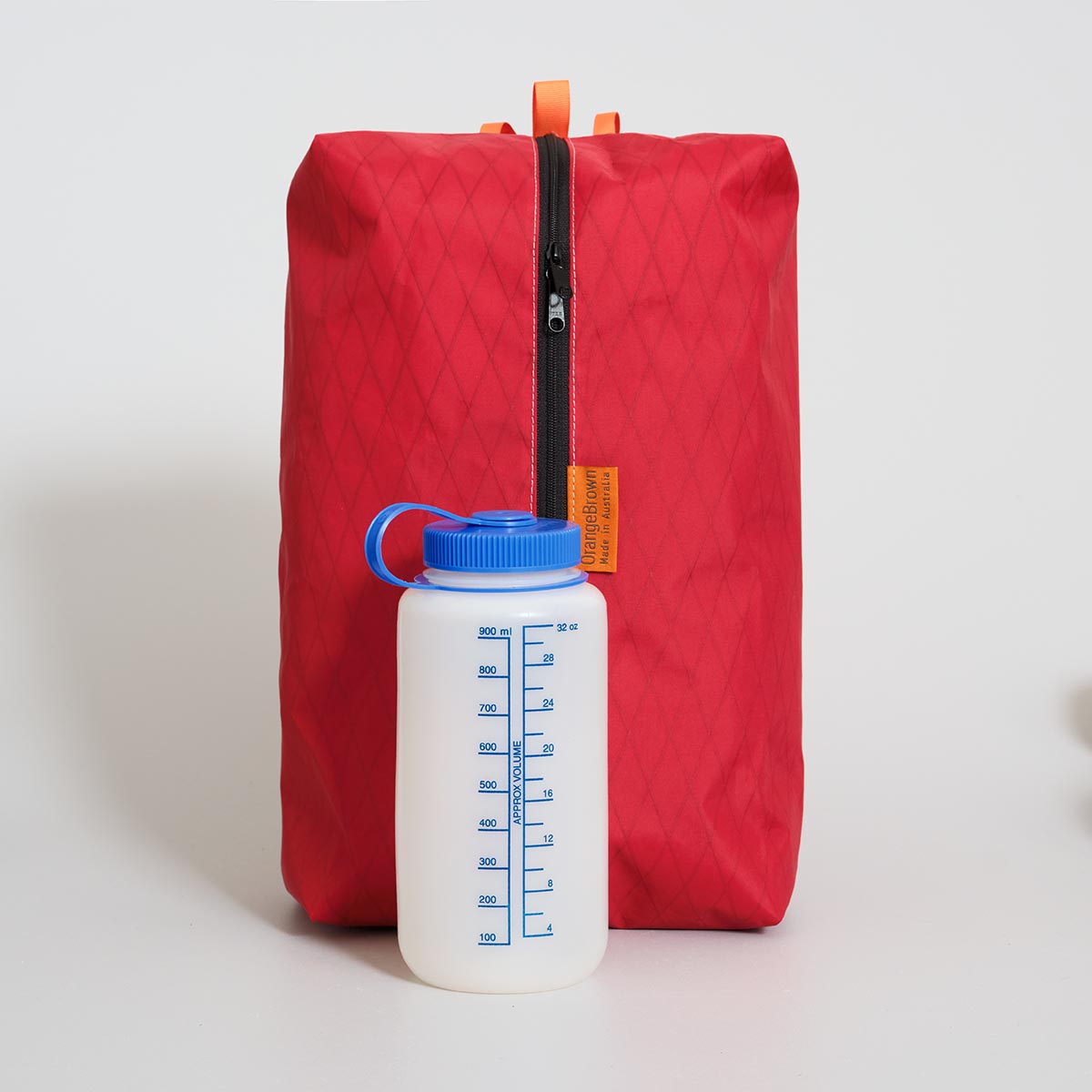 Red large packing cube with a black zipper made with X-Pac VX21 fabric on a light gray background. A white water bottle with blue cap placed in front for size comparison