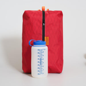 Red large packing cube with a black zipper made with X-Pac VX21 fabric on a light gray background. A white water bottle with blue cap placed in front for size comparison