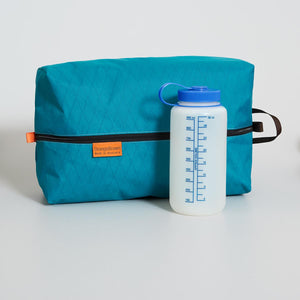 Teal packing cube with black zipper and orange brand label next to a white water bottle with blue cap on a light gray background